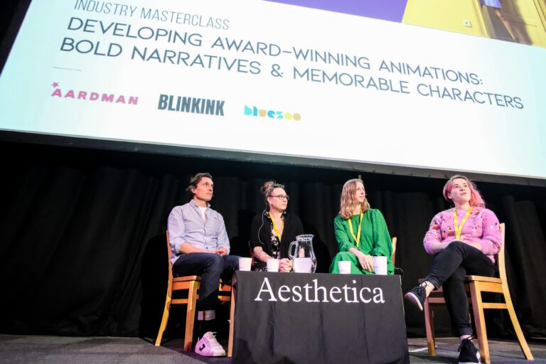 4 panelists sat on a stage with a table and cloth saying aesthetica on it. A projector screen is behind them. They are all using headset microphones.